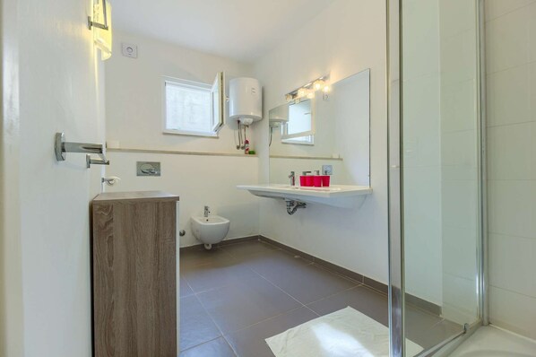 A modern bathroom with a wooden vanity, a pedestal sink, a bidet, and a glass shower enclosure.