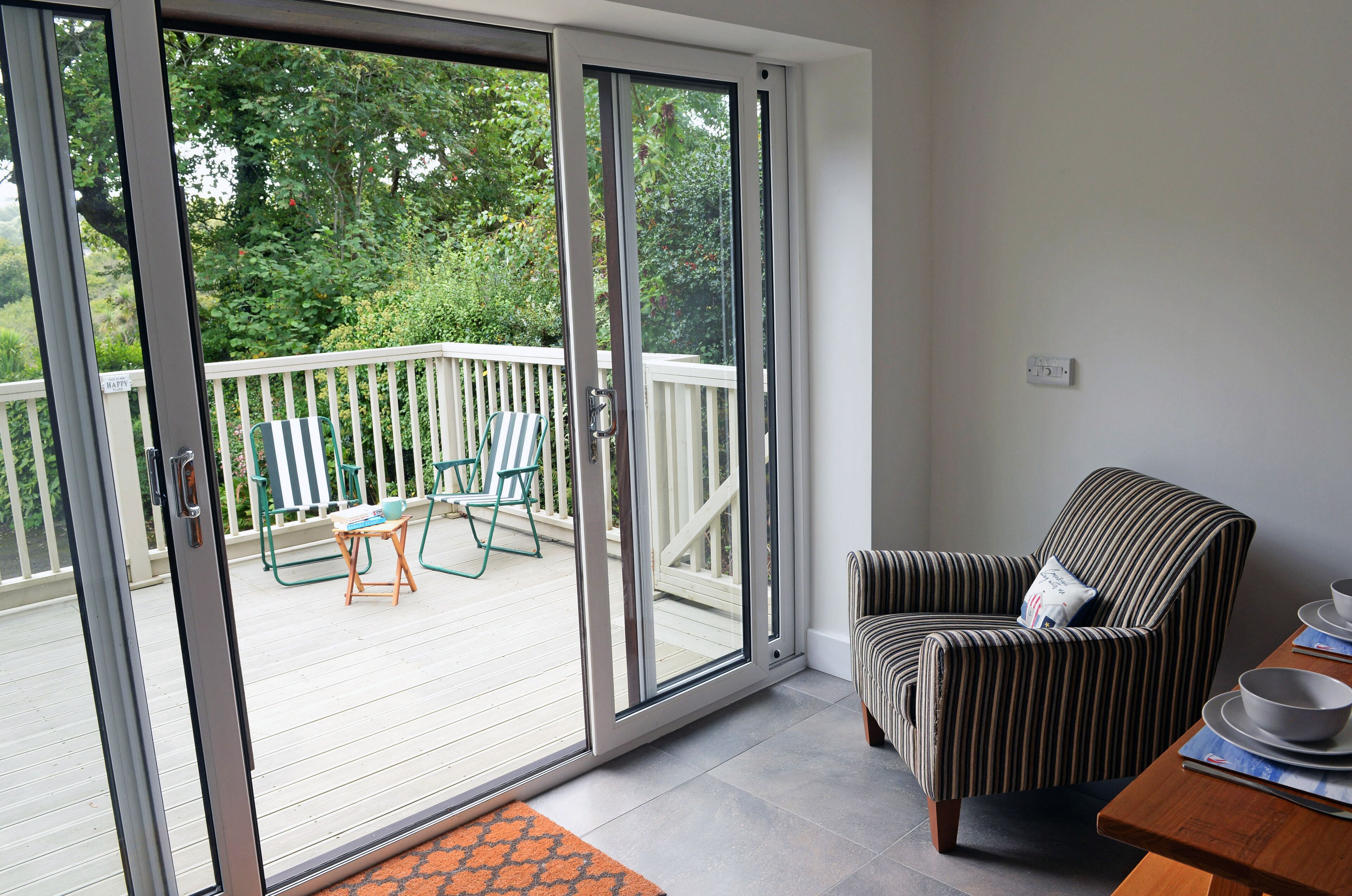 Ein Zimmer mit einer Schiebetür zu einer Terrasse, ein gestreifter Sessel und ein Esstisch mit Tellern und Schüsseln.