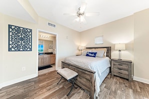 Quarto com cama de madeira, mesa de cabeceira, um banco, ventilador de teto e vista para a cozinha.
