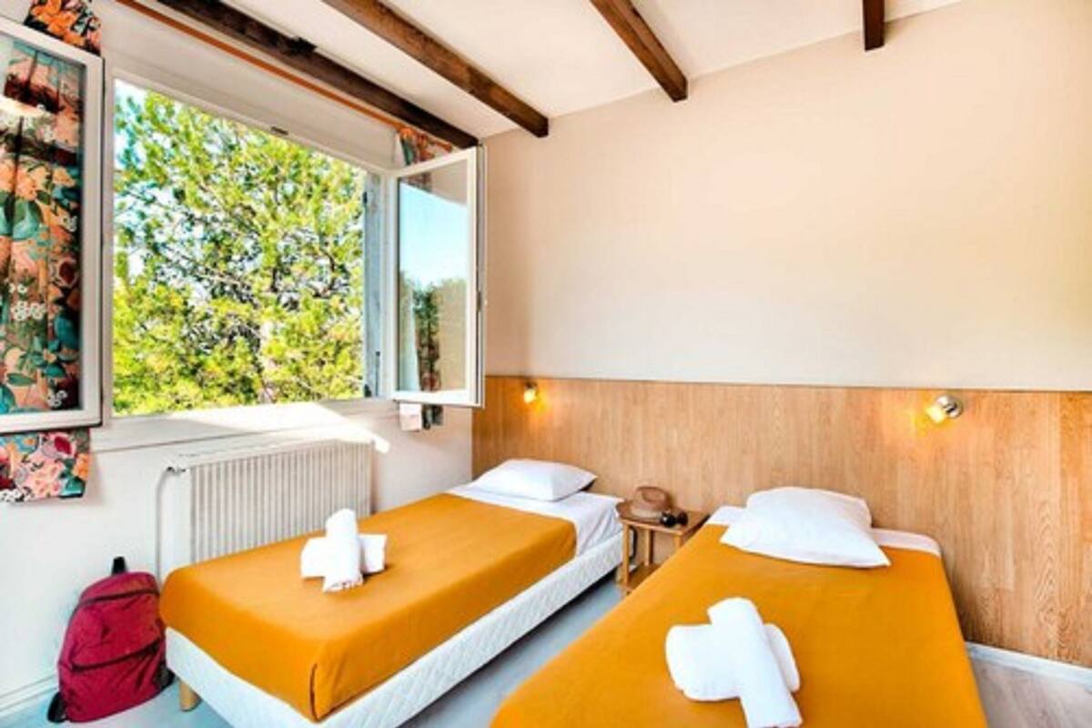 Habitación con dos camas, cada una con una almohada blanca y una toalla. Se ve una ventana con vistas a árboles, un radiador y una mochila roja en el suelo.