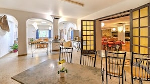 Un vaste espace dédié à la salle à manger, avec une table et des chaises en marbre, une plante en pot, et une vue sur une autre pièce où une table est dressée pour le repas.
