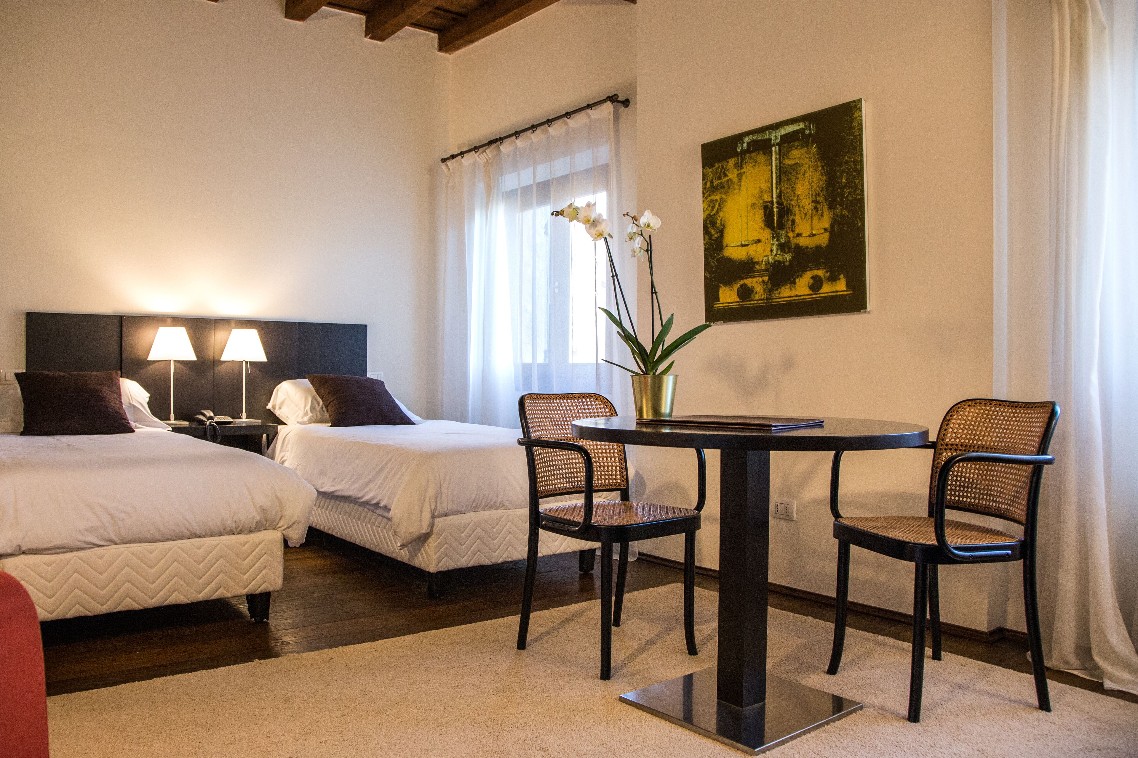 A hotel room with two beds, a dining table with chairs, a vase with flowers, and a painting on the wall.