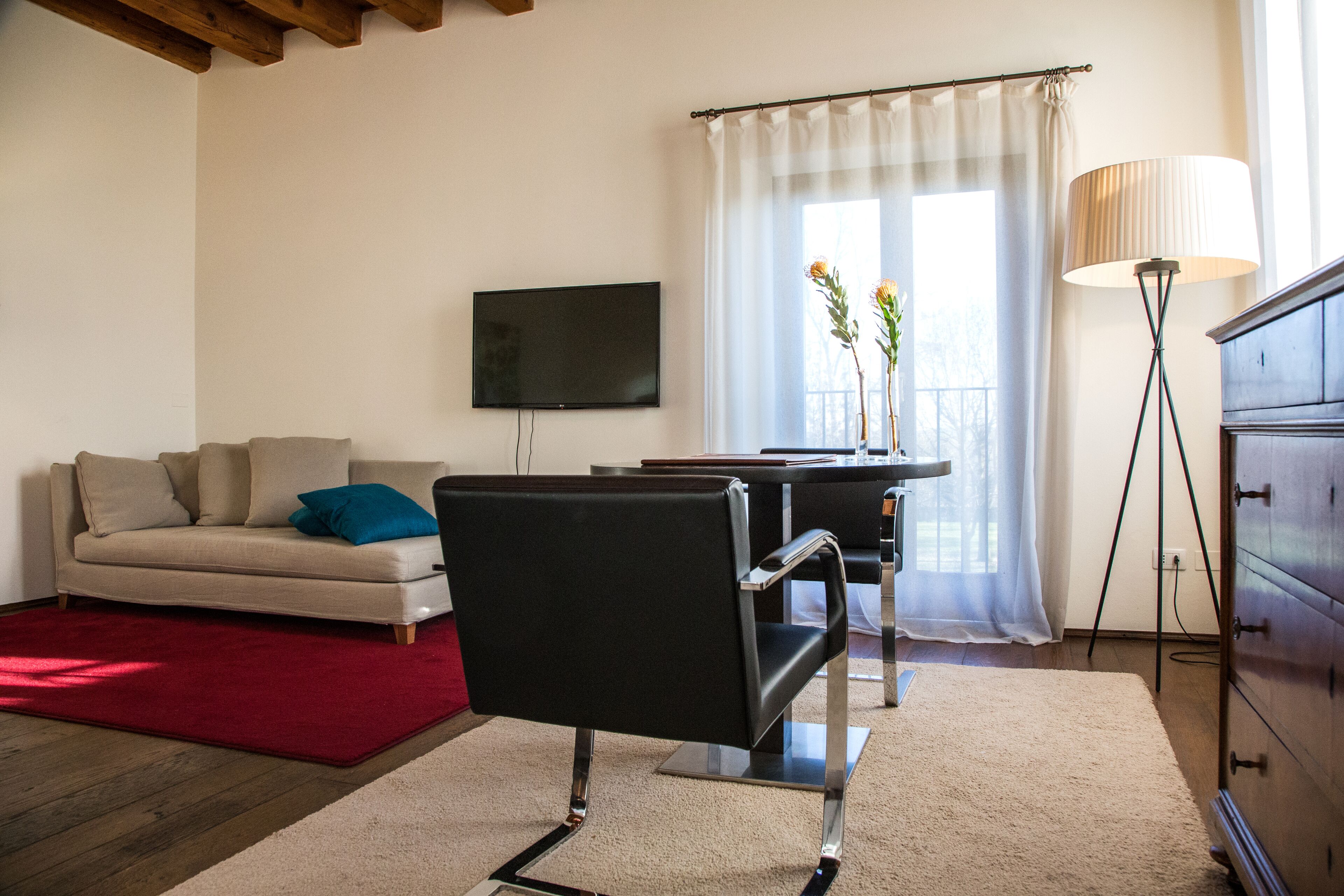 Una sala moderna con un sofá, una silla negra, un televisor, una lámpara y un ventanal con cortinas.
