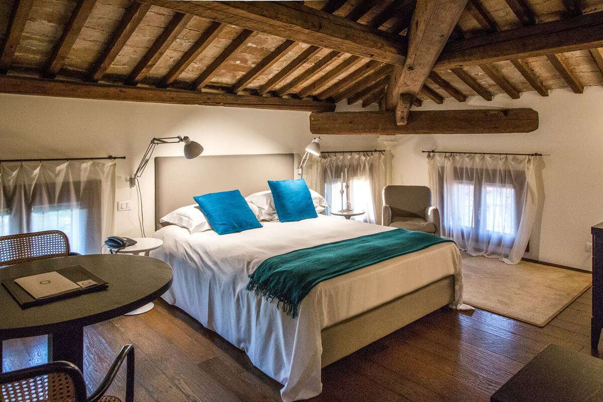 A bedroom with a wooden ceiling, a bed with blue pillows, a bedside table, a chair, and a desk with a lamp.
