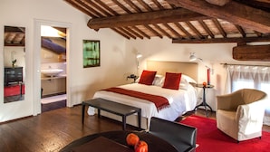 A bedroom with a wooden ceiling, a bed with white bedding and red pillows, a bedside table with a lamp, a chair, and a small table with fruits. - Relais Villa Ormaneto (Cerea)