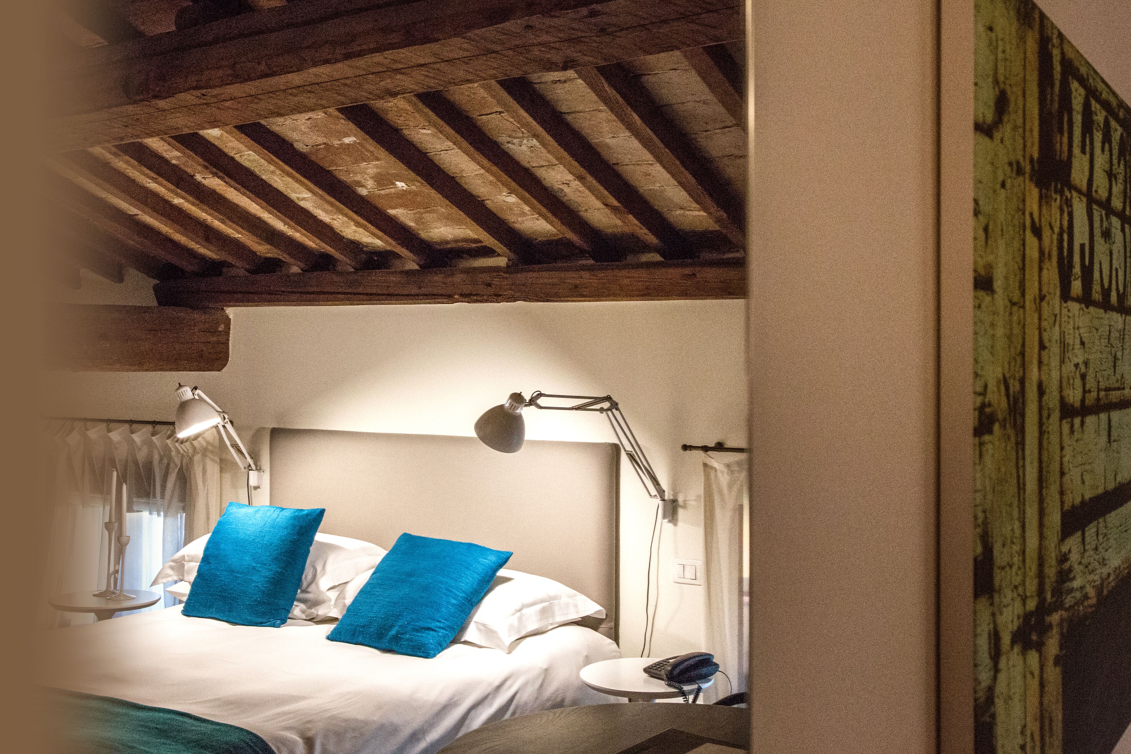 A hotel room with a wooden ceiling, a bed with white linens and two blue pillows, a bedside table with a lamp, and a telephone.