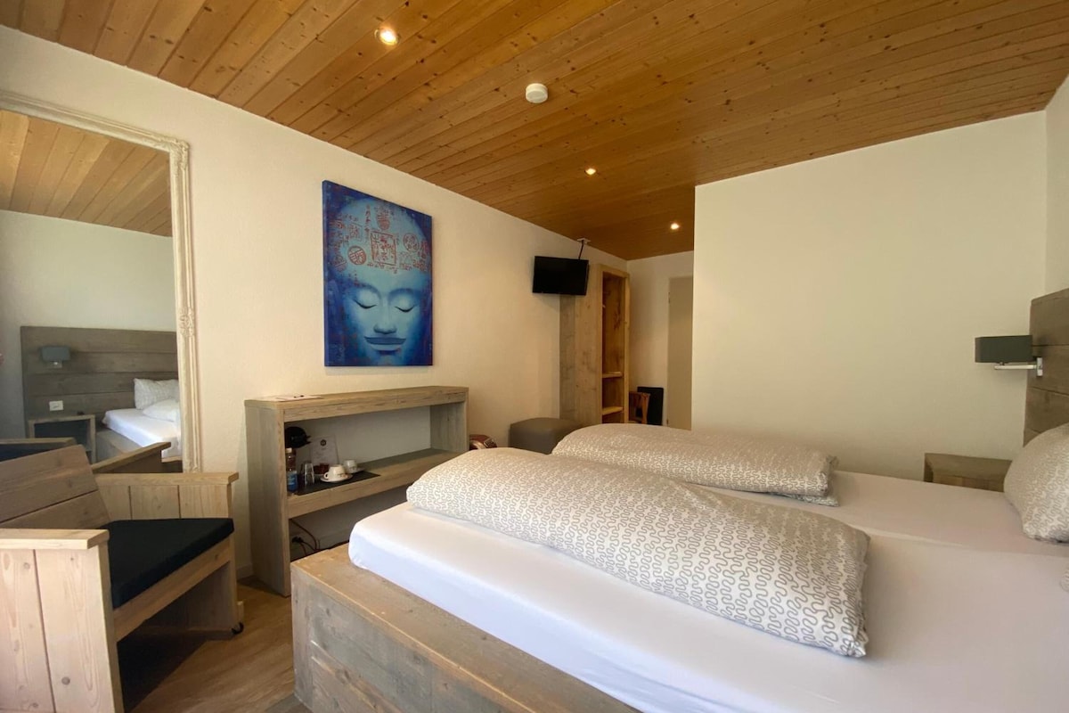A bedroom with a wooden ceiling, a bed with patterned bedding, a bedside table, a small wooden bench, a wall-mounted TV, and a painting on the wall.