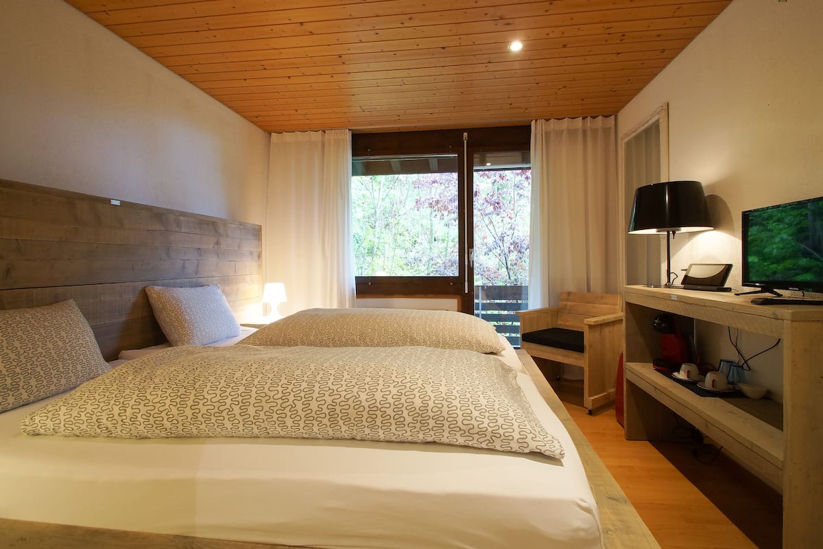 A bedroom with a wooden ceiling, a bed with patterned bedding, a desk with a computer, and a TV.