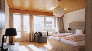 A bedroom with a wooden ceiling, a bed with pink bedding, a grey sofa, and a large window with curtains. - Hotel Twing (Hasliberg)