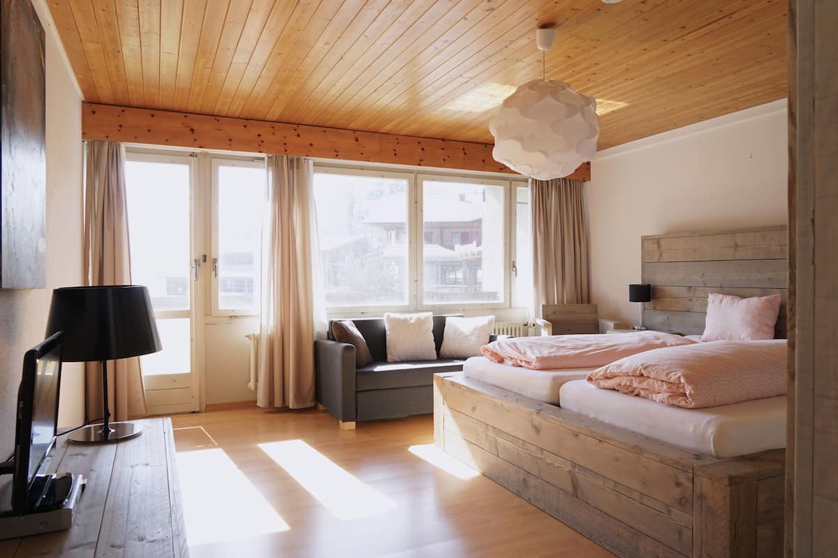 A bedroom with a wooden ceiling, a bed with pink bedding, a grey sofa, and a large window with curtains.