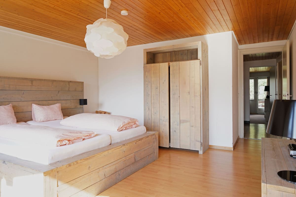 A bedroom with a wooden headboard, a large bed with pink bedding, a wooden wardrobe, and a dining area with a table and chairs.