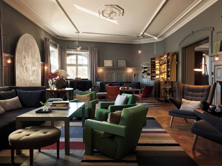 A spacious living room with a sofa, armchairs, a coffee table, and a fireplace. There is a bookshelf filled with books and decorative items.