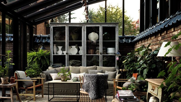 A cozy patio area with a cushioned seating arrangement, a wooden table, and a decorative cabinet filled with vases. - Ett Hem (Stockholm)