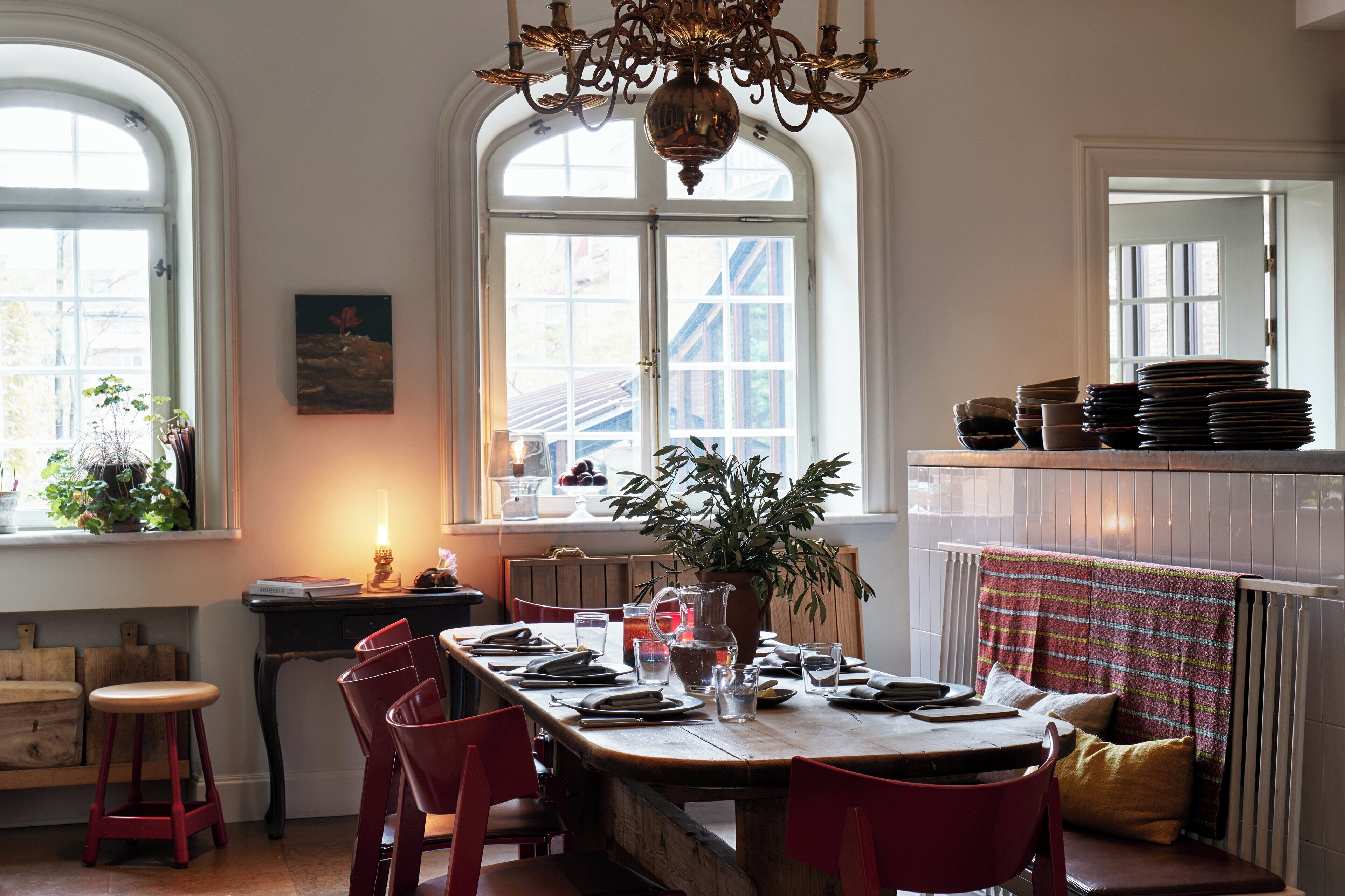 A dining area with a long table set for four, surrounded by red chairs, a potted plant, and a chandelier.