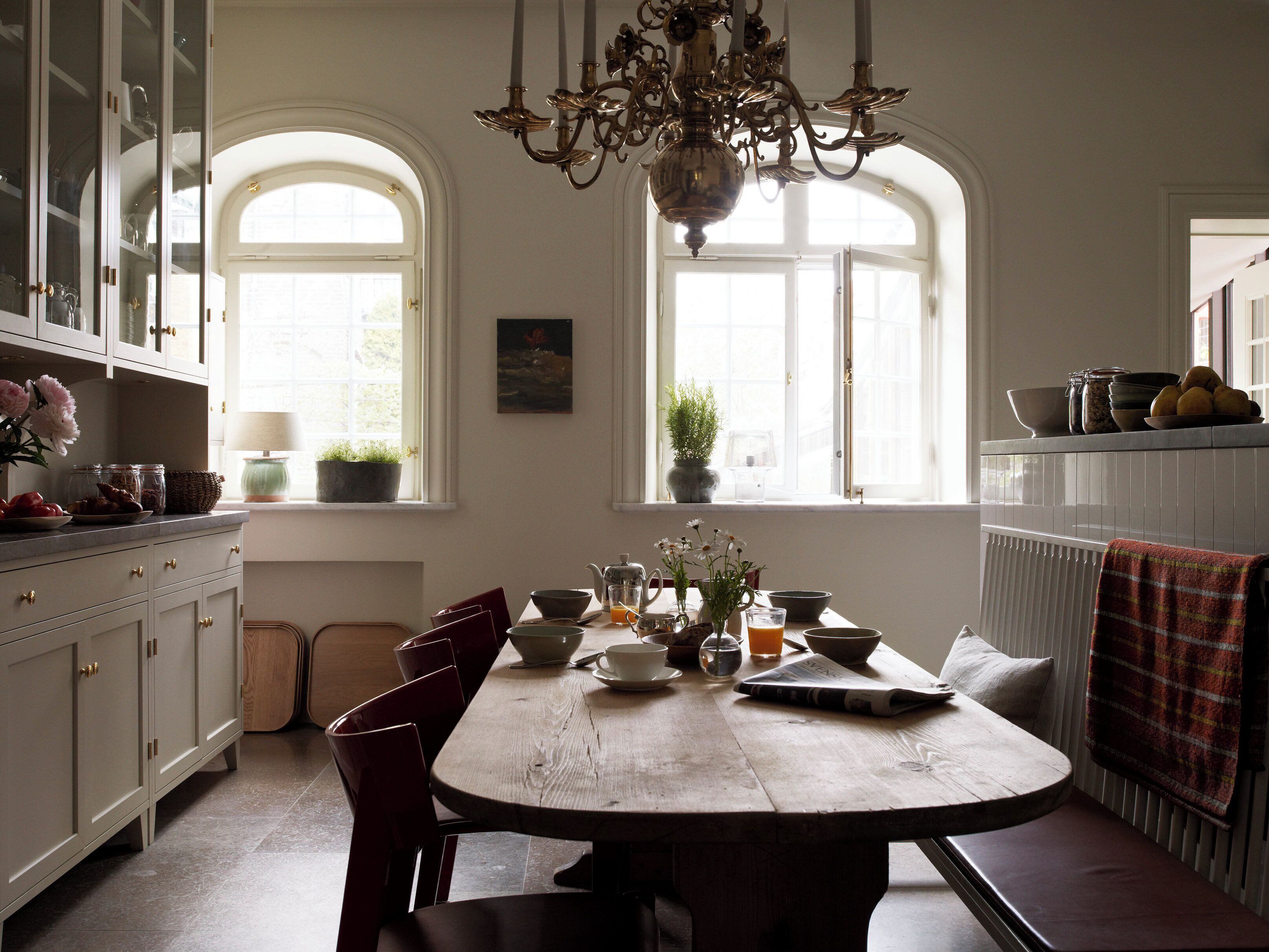 Un comedor con una mesa de madera, sillas y una araña. Se observan ventanas con arcos y un radiador en la pared.