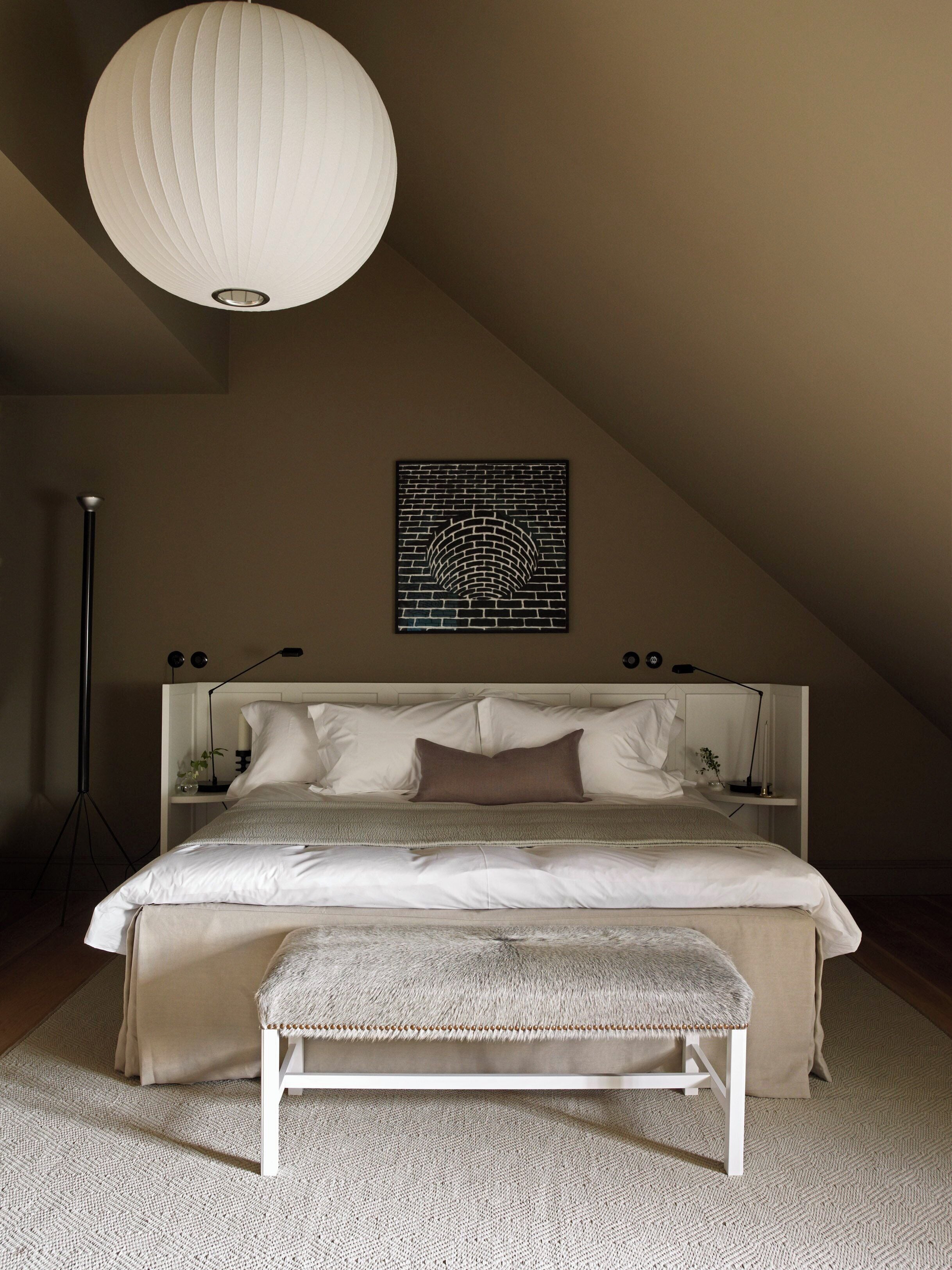 A bedroom with a large bed, a bench, a hanging light fixture, and a wall-mounted artwork.