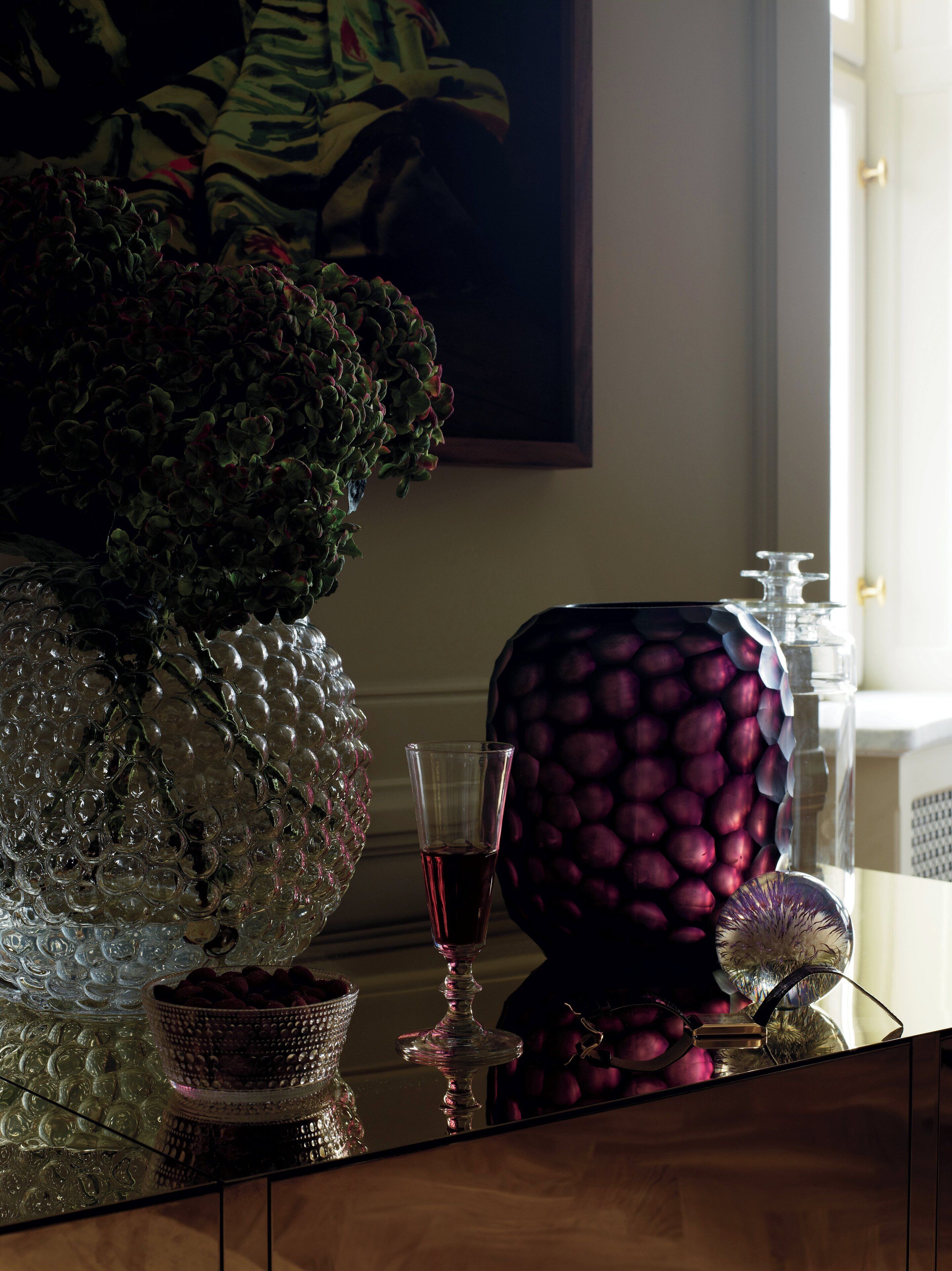 Una mesa con un jarrón de vidrio decorativo, una copa con líquido rojo y un recipiente estampado.