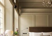 A bedroom with a large bed, a bedside table with a lamp, a chair, and a desk with a lamp and books.
