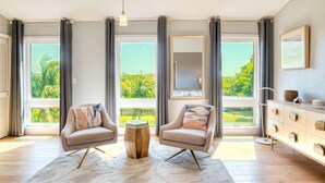Una sala de estar moderna con dos sillones, una mesita de centro y un mueble de madera. Amplios ventanales ofrecen vistas al verde exterior.