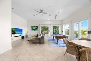 A spacious living area with a pool table, comfortable seating, a flat-screen TV, and a large window offering a view of the outdoors.