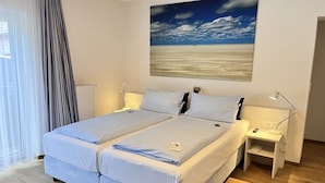 Ein Doppelbett mit Kopfteil, Nachttische mit Lampen, ein Gemälde mit Strandmotiv und ein Fenster mit Vorhängen.