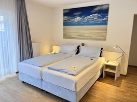 Ein Doppelbett mit Kopfteil, Nachttische mit Lampen, ein Gemälde mit Strandmotiv und ein Fenster mit Vorhängen.