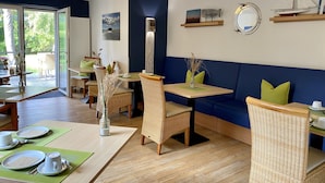 Ein Restaurant mit Holztischen, grünen Kissen und Blick durch große Fenster.