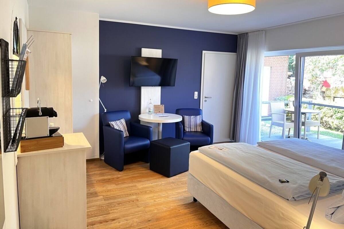 A modern hotel room with a bed, a desk, two armchairs, a TV, and a balcony with outdoor seating.