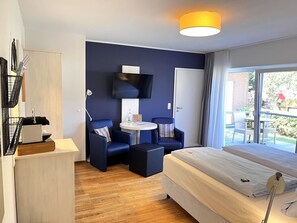 A modern hotel room with a bed, a desk, two armchairs, a TV, and a balcony with outdoor seating. - Hotel garni DünenNest (St. Peter-Ording)