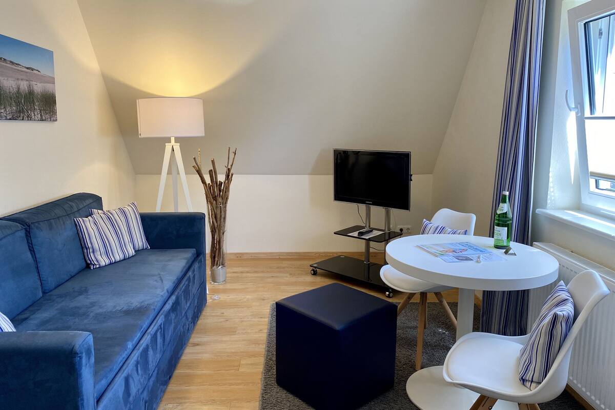 A modern room with a blue sofa, a small round table with chairs, a flat-screen TV, a lamp, and a window with curtains.