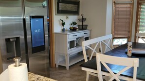 Et køkken med et rustfrit stål køleskab, en mikroovn, kaffemaskine, en hvid bordplade, og et spiseområde med en hvid stol og et bord.