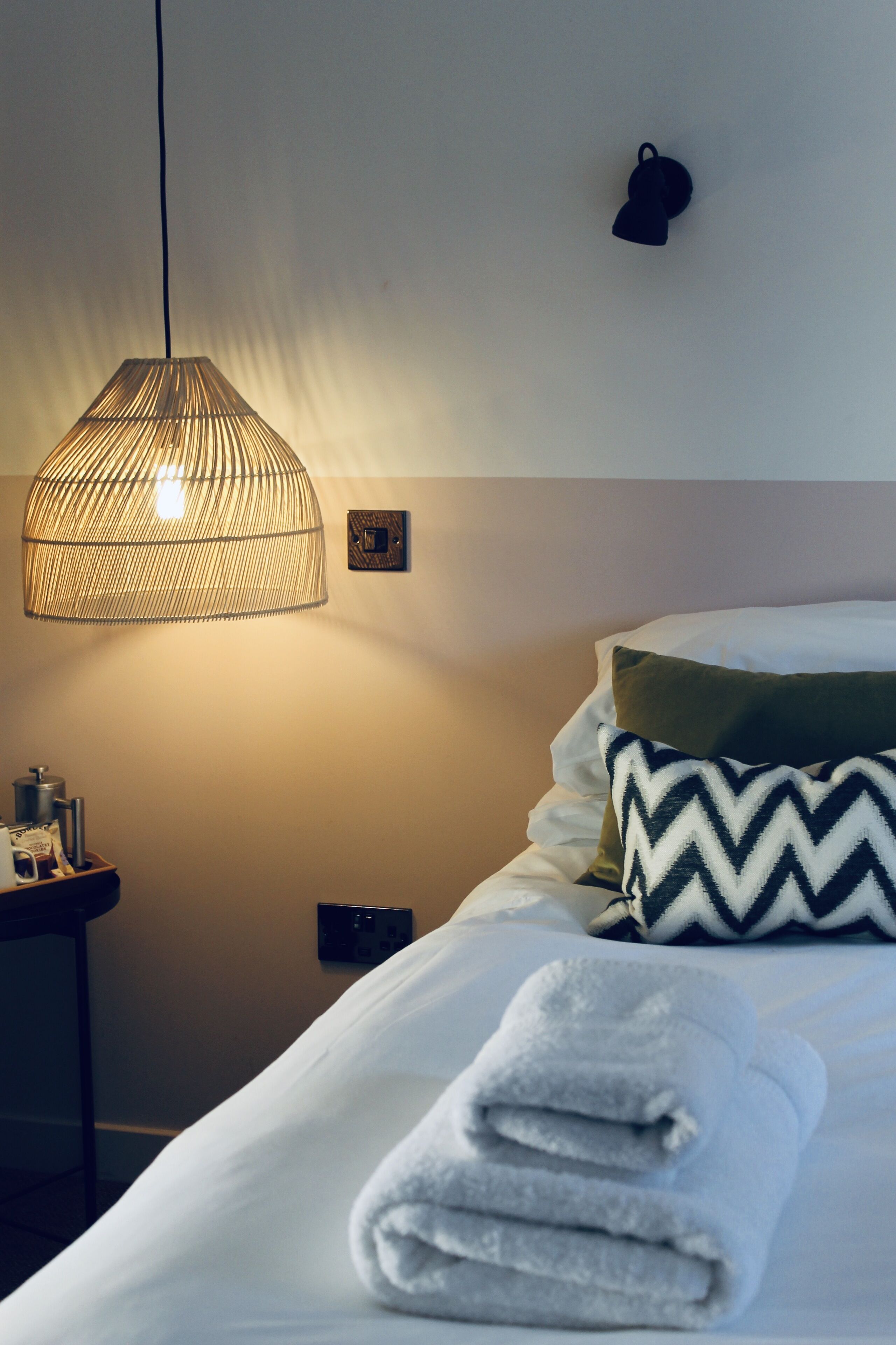 A neatly made bed with a folded towel, a hanging light fixture, and a wall-mounted light switch.
