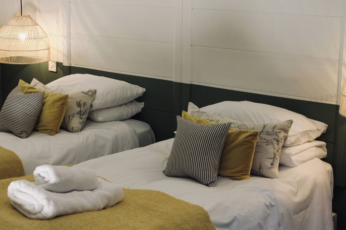 Ein Hotelzimmer mit zwei Betten, beide mit weißen Leintüchern und mehreren Kissen. Über den Betten befinden sich je eine Hängelampe.