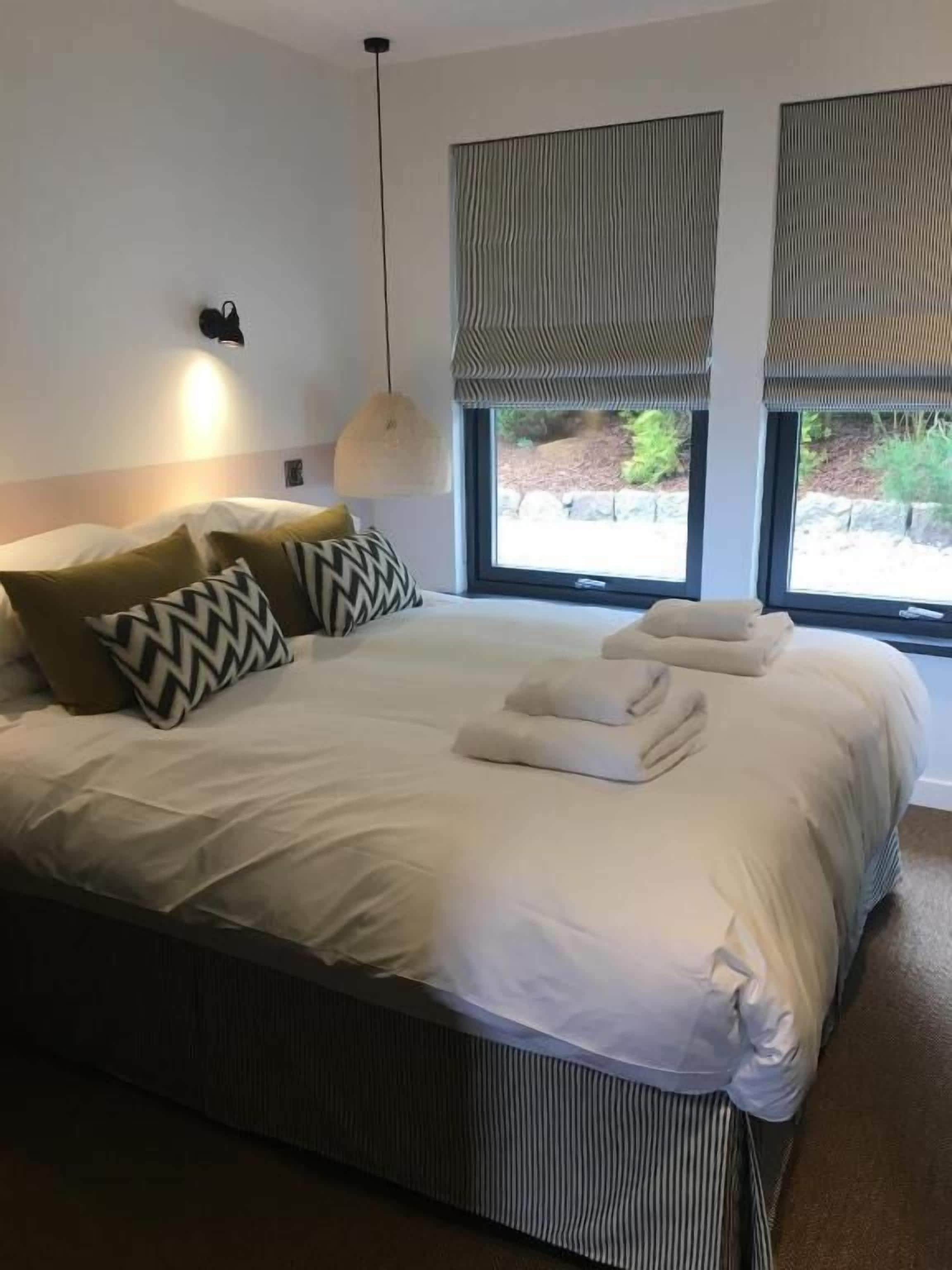 A neatly made bed with pillows, a hanging light fixture, and large windows with blinds.