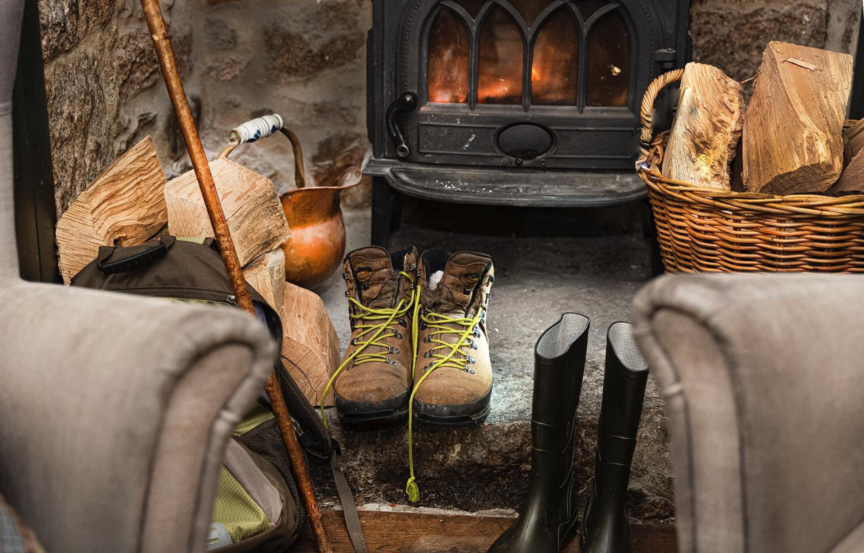 Una habitación acogedora con chimenea, leña, un cesto de astillas y un par de botas junto a un bastón de senderismo.