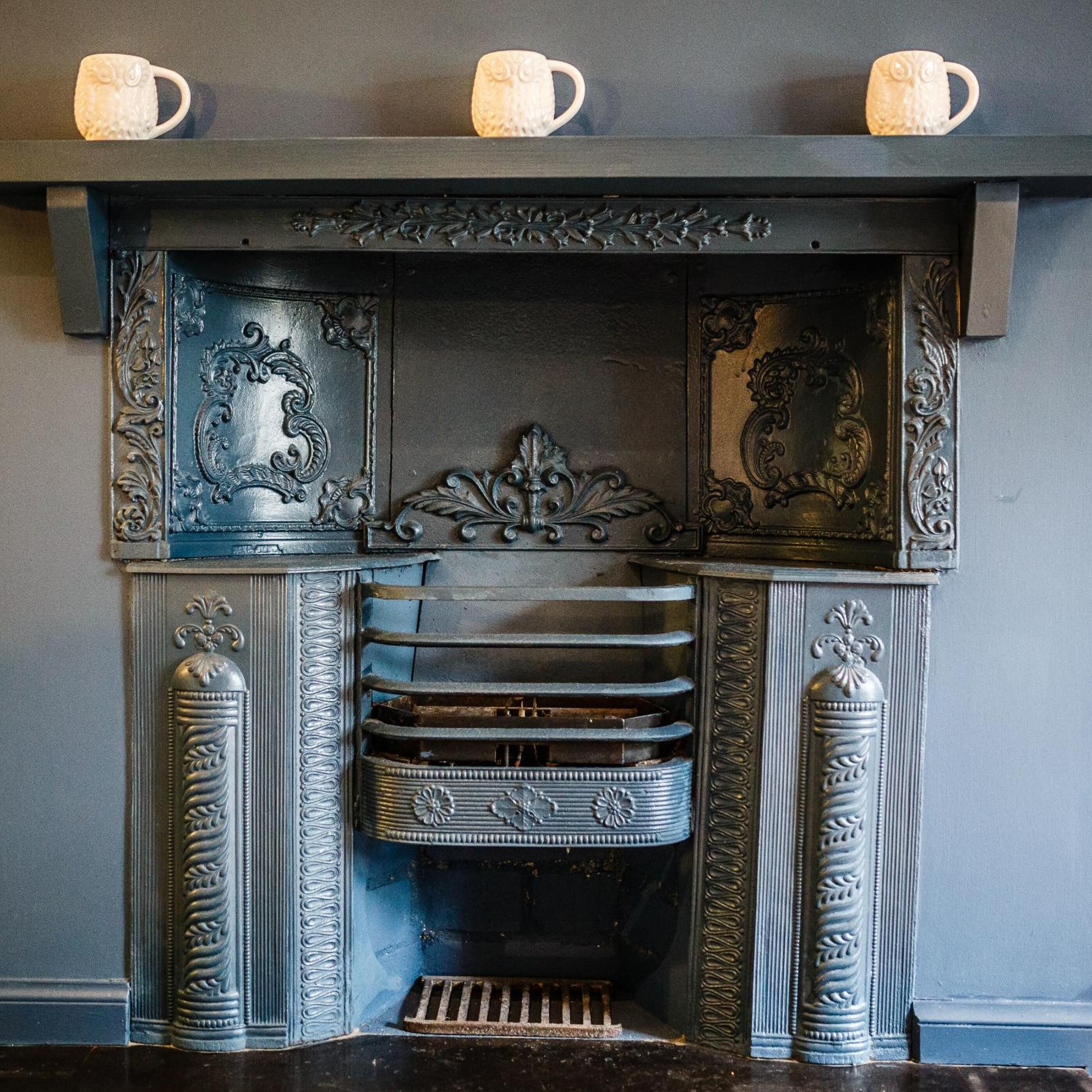 A fireplace with ornate metal detailing and three white ceramic cups on the mantel.