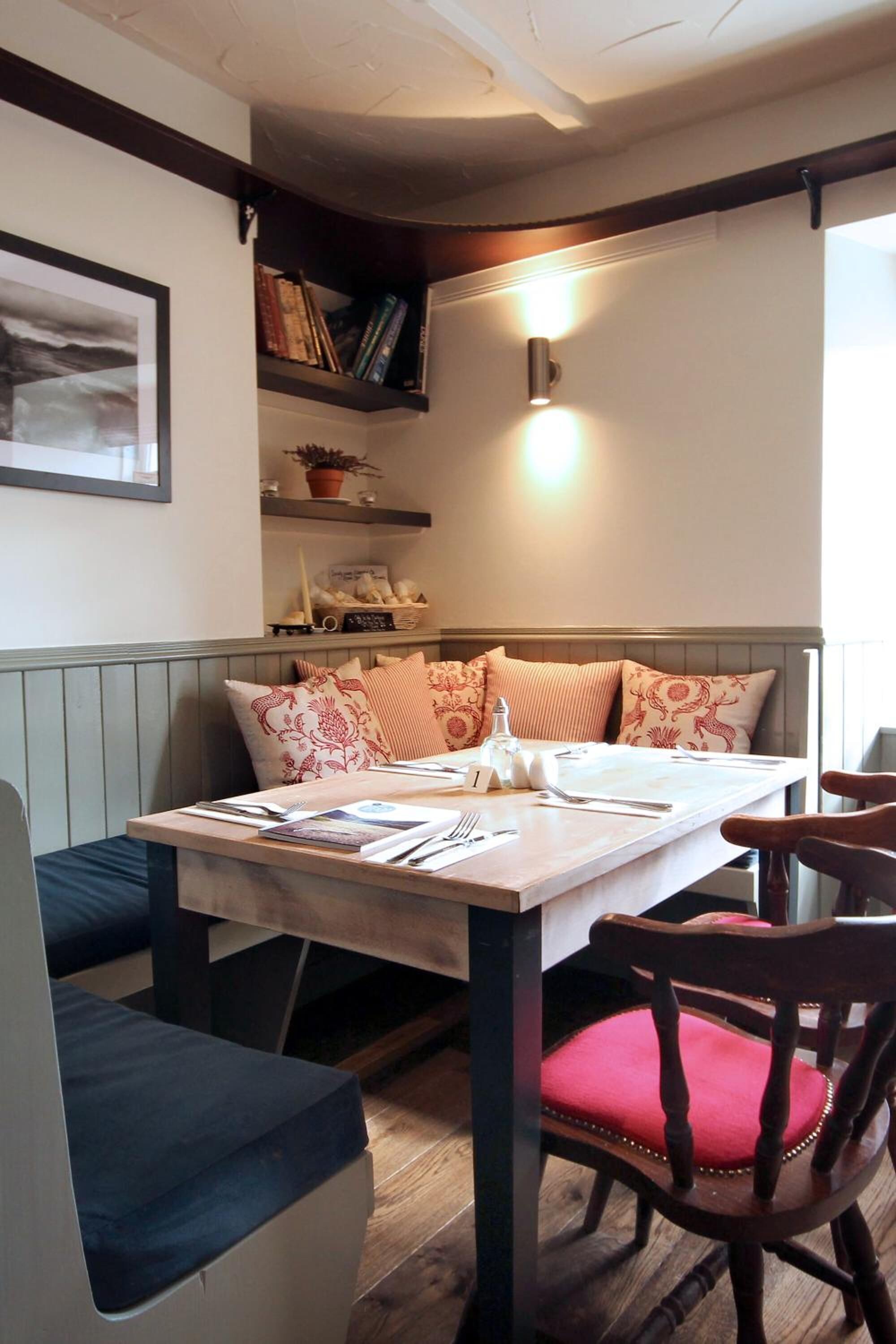 Un coin repas avec une table en bois dressée pour deux personnes, un banc agrémenté de coussins et une bibliothèque contenant des livres et des objets décoratifs.