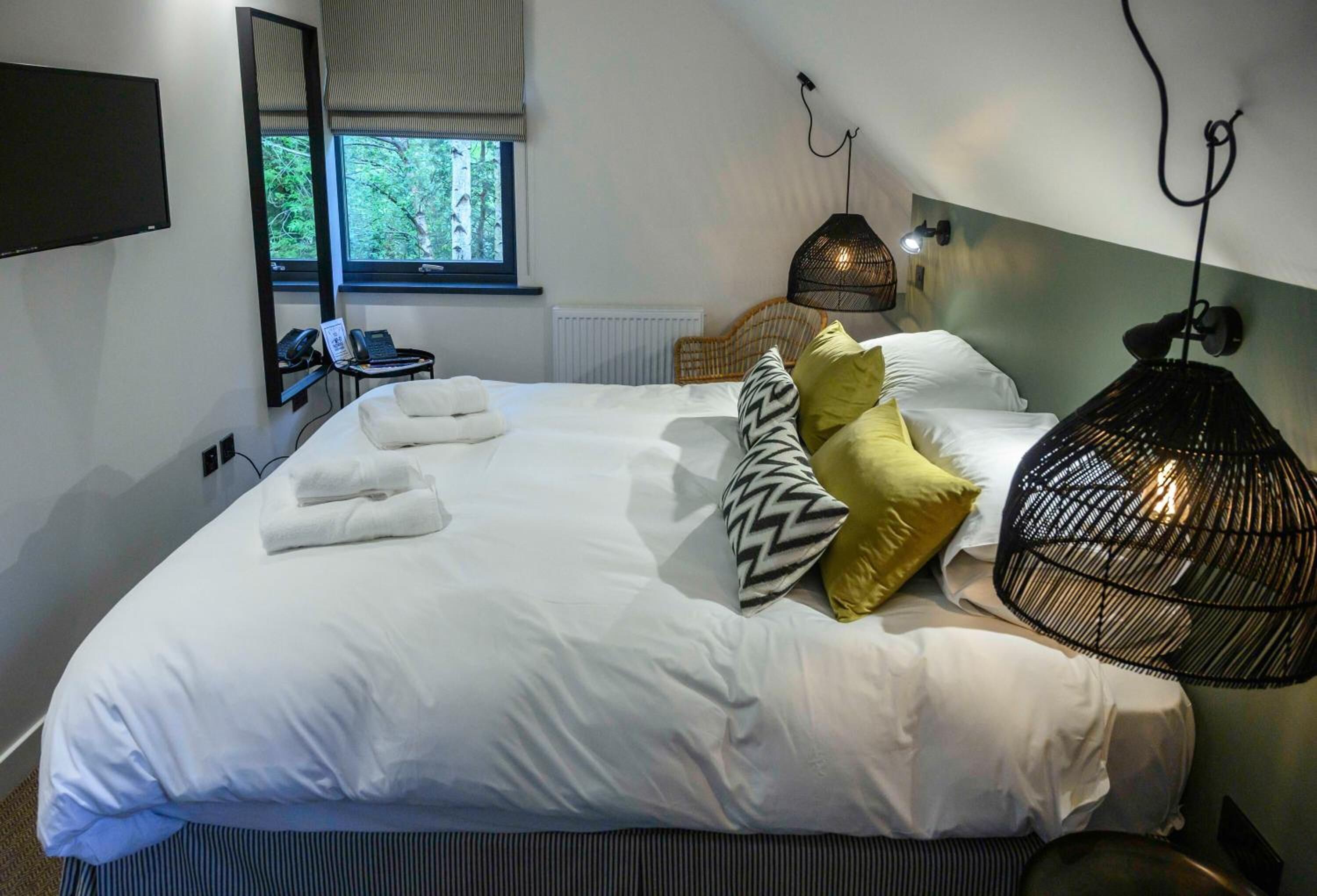 A neatly made bed with white linens, a bedside table with a lamp, and a window with a view of trees.