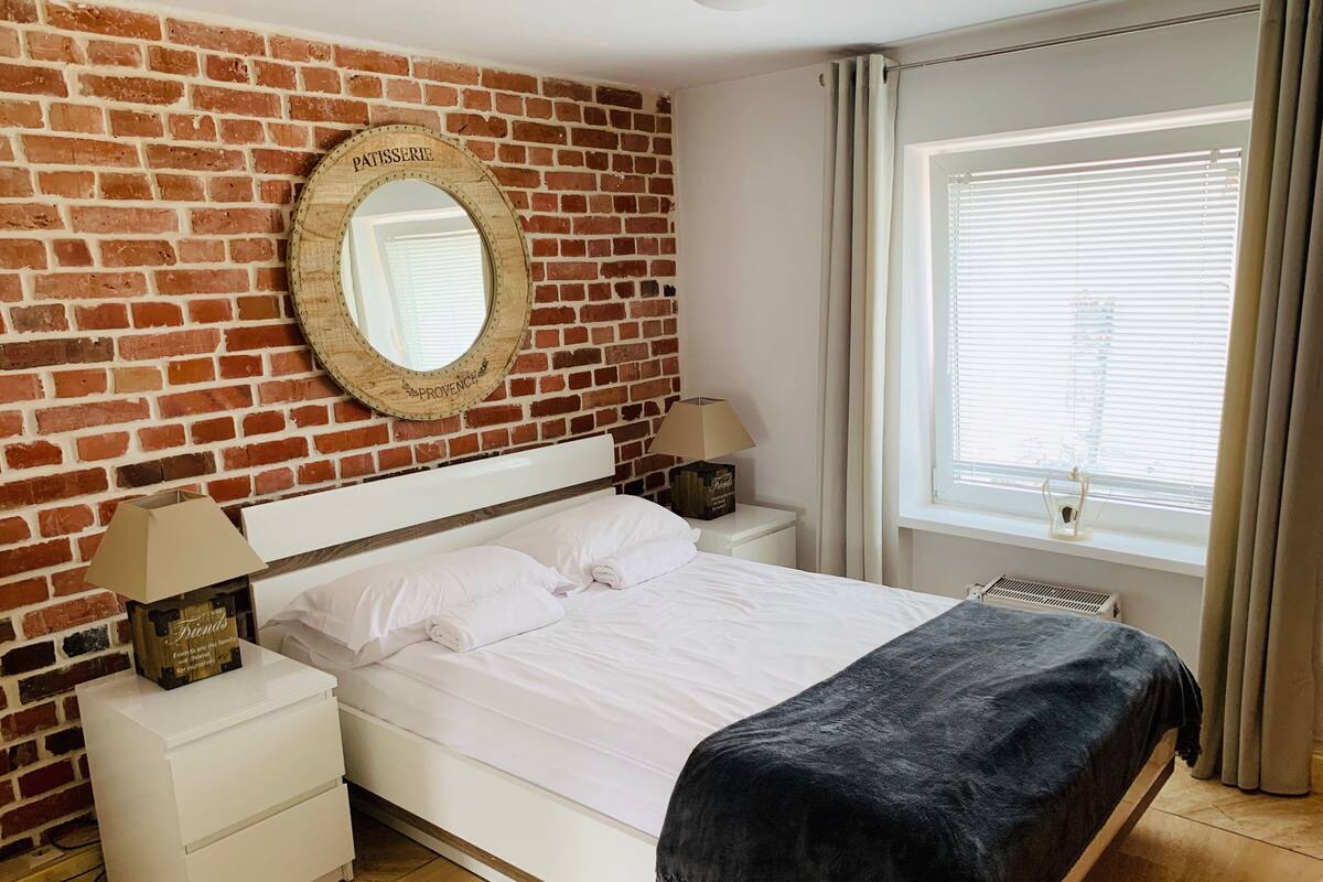 Un dormitorio con pared de ladrillo, una cama con ropa de cama blanca, dos mesitas de noche con lámparas, una ventana con persianas y un espejo con la palabra 'PATTISSEERIE' en la pared.