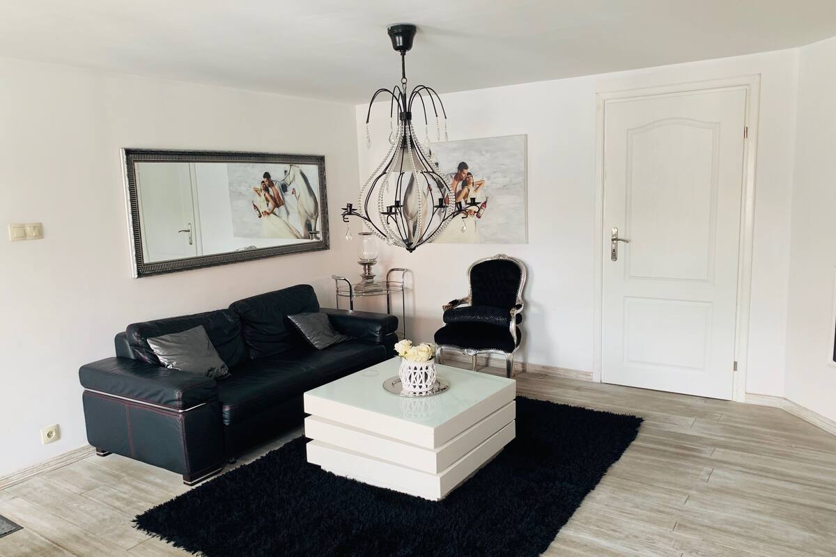Un salón moderno con un sofá de cuero negro, una mesa de centro blanca, una alfombra negra, una araña y una puerta blanca.