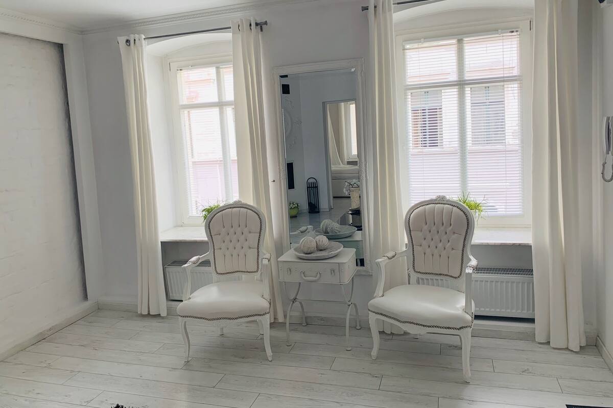 Una habitación con dos sillones blancos tapizados, un tocador con espejo y ventanas amplias con cortinas blancas.