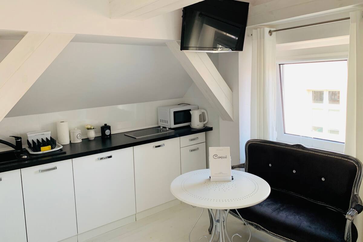 Cocina compacta con gabinetes blancos, encimera negra, microondas, cafetera y una pequeña mesa con una silla.