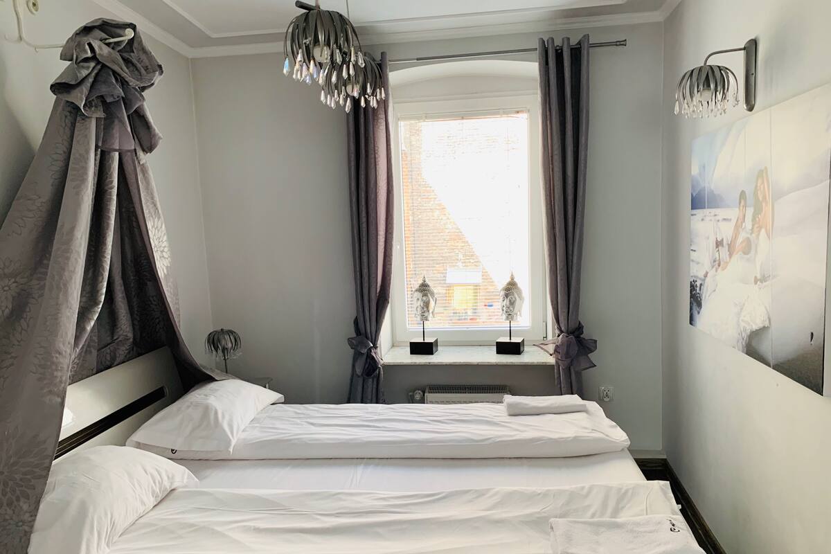 Un dormitorio con cama, ventana con cortinas, lámpara colgante y un cuadro en la pared.