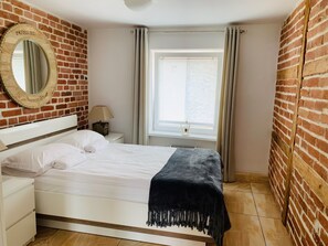 A bedroom with a brick wall, a round mirror, a bed with white bedding, a nightstand, a lamp, and a window with curtains. - Apartamenty Campari (Torun)
