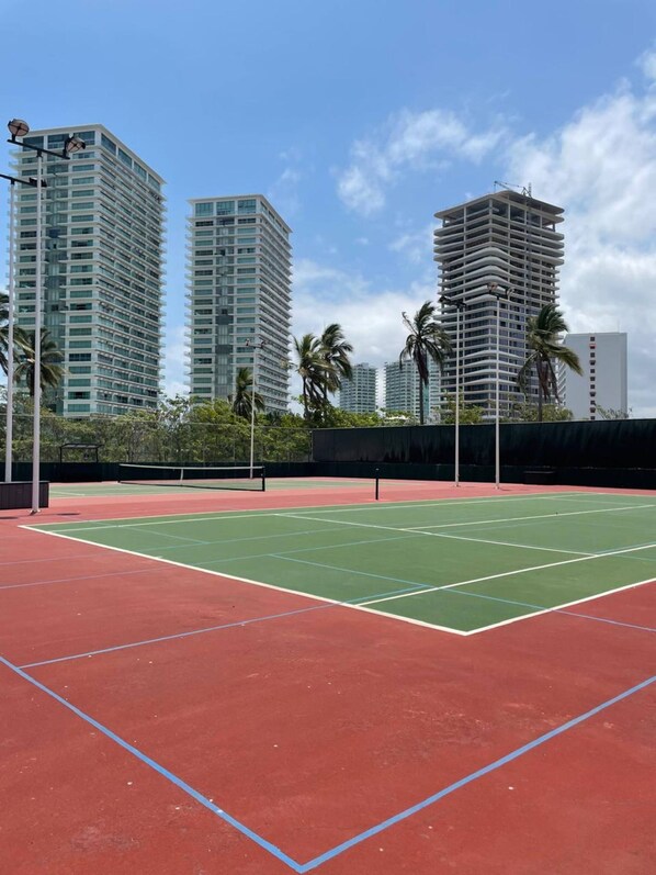 A tennis court with a net, surrounded by tall buildings and palm trees.