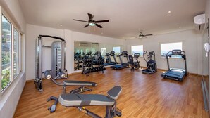 Une salle de sport spacieuse, équipée de divers appareils d’exercice, dotée de grandes fenêtres et d’un ventilateur au plafond.