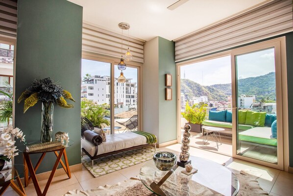 Una sala de estar moderna con un sofá, una mesa de centro de vidrio y vistas a un paisaje urbano a través de amplios ventanales.