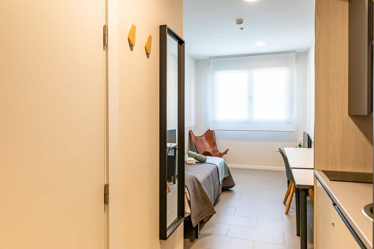 A modern bedroom with a large mirror, a bed with a brown headboard, a desk, and a window providing natural light.