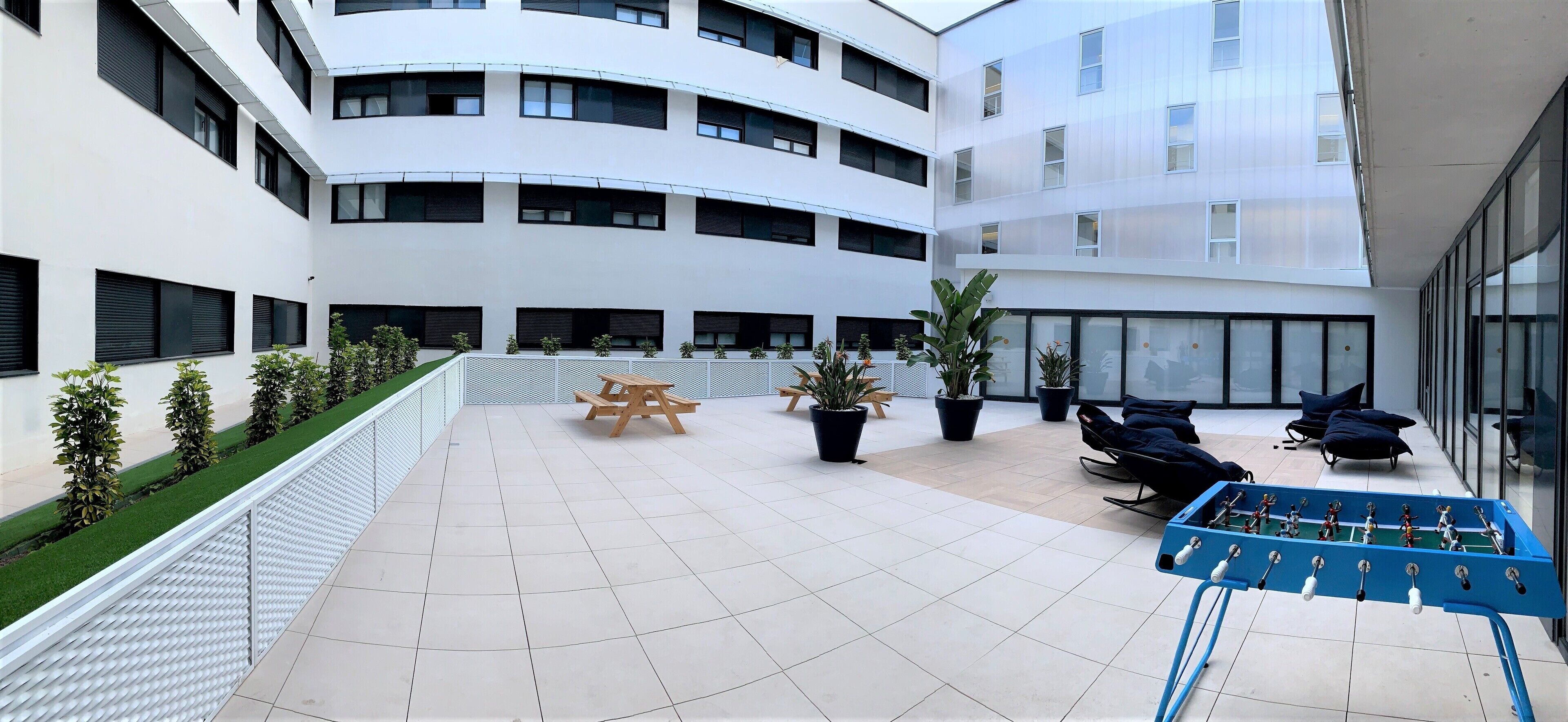 Un edificio moderno de varios pisos, un patio con piso de baldosas, plantas en macetas, un banco de madera y una mesa de fútbol de mesa.