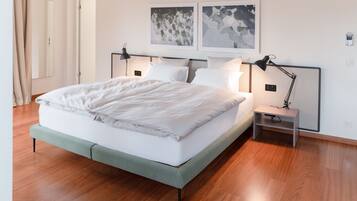 Een net opgemaakt bed met witte lakens, een nachtkastje met een lamp, en twee ingelijste foto's aan de muur.